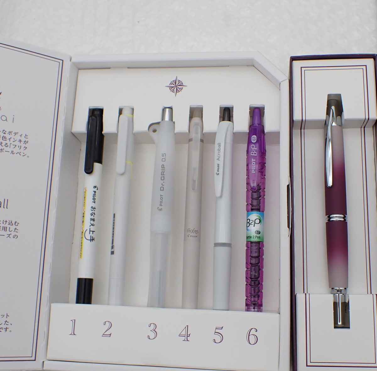 白い箱に並んだボールペン6本と、となりの箱に入った紫色の万年筆